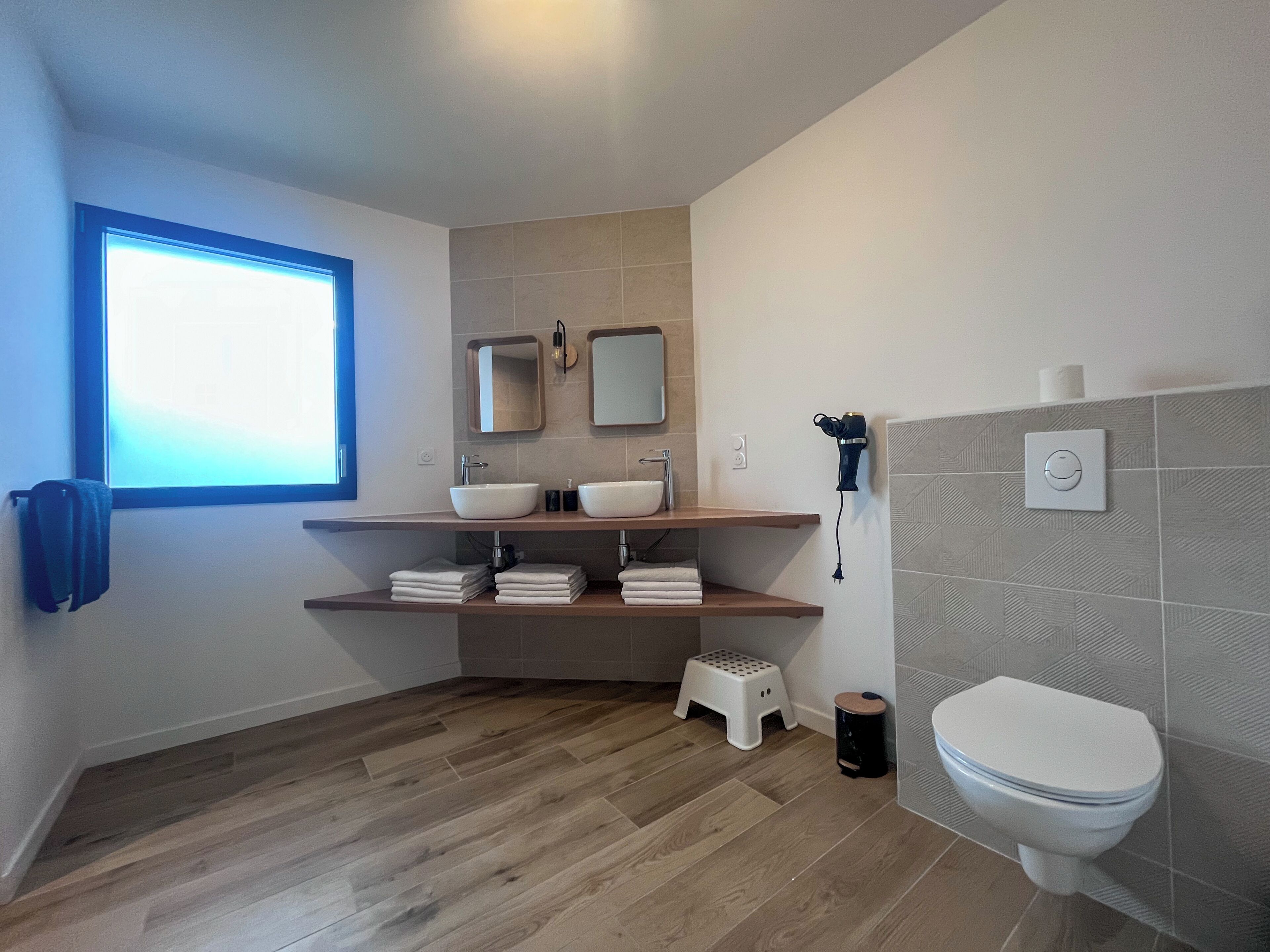 A modern bathroom with a window, two sinks, a toilet, and a hairdryer.