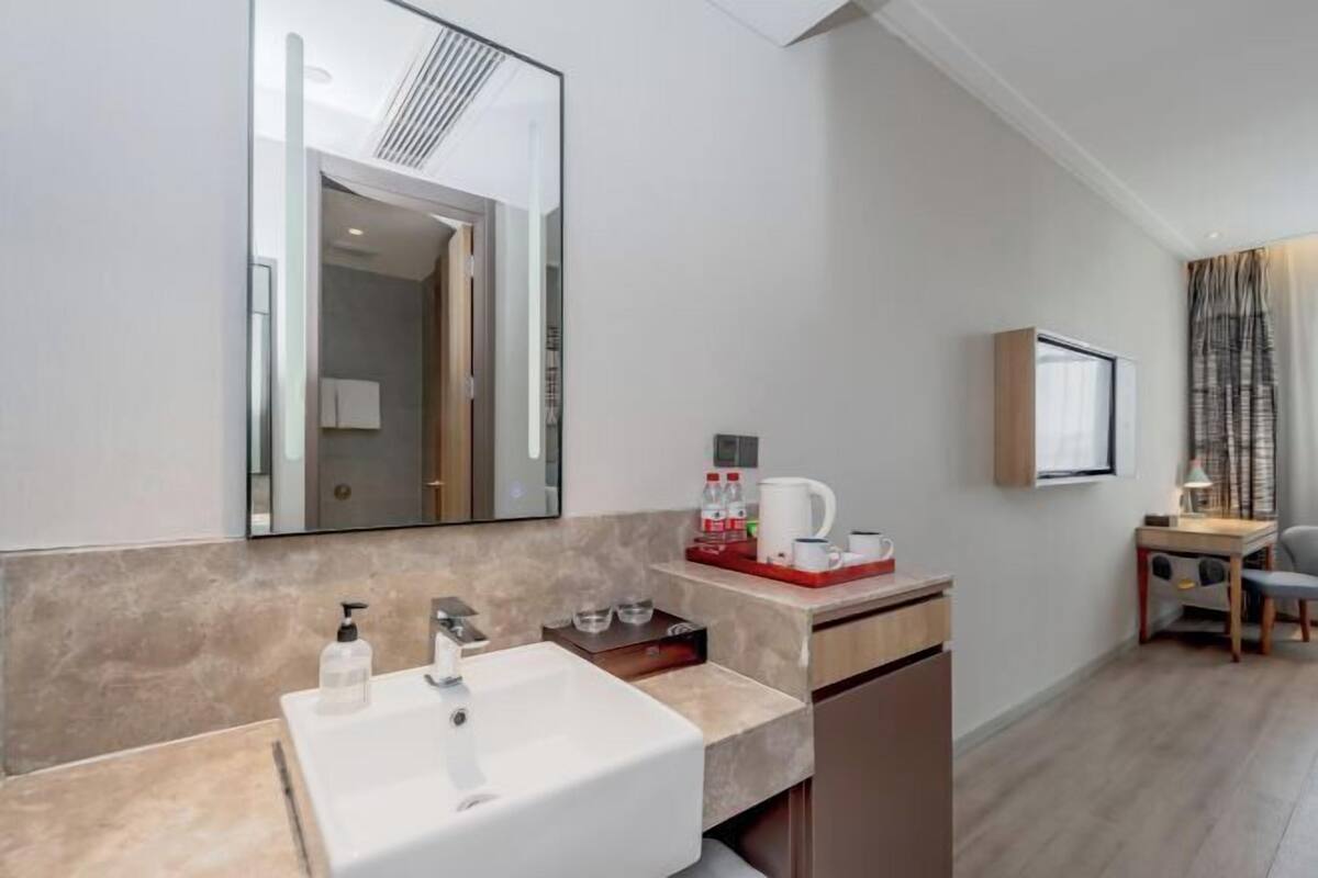 A modern bathroom with a large mirror, a white sink, and a small cabinet. There is a wooden table with a lamp and a chair in the adjacent room.
