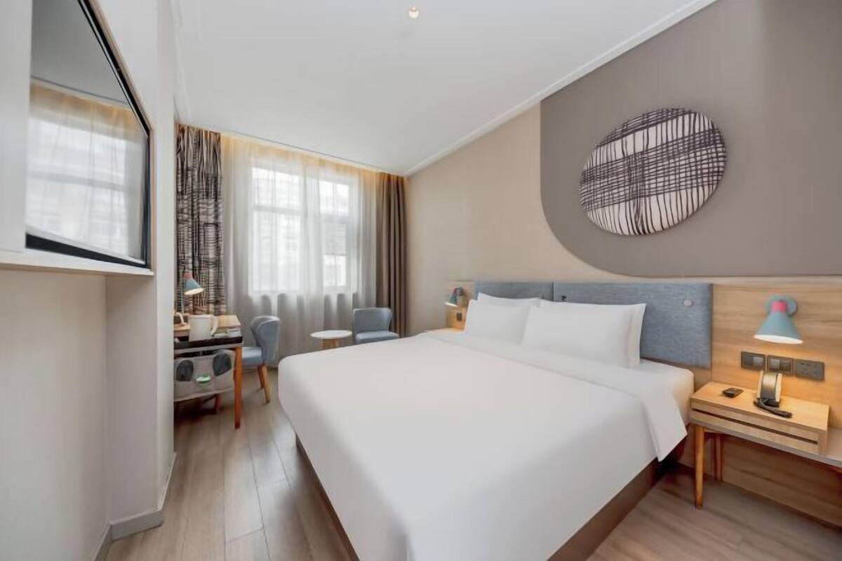 A hotel room with a large bed, a desk with a lamp, a chair, a round wall decoration, and a window with curtains.