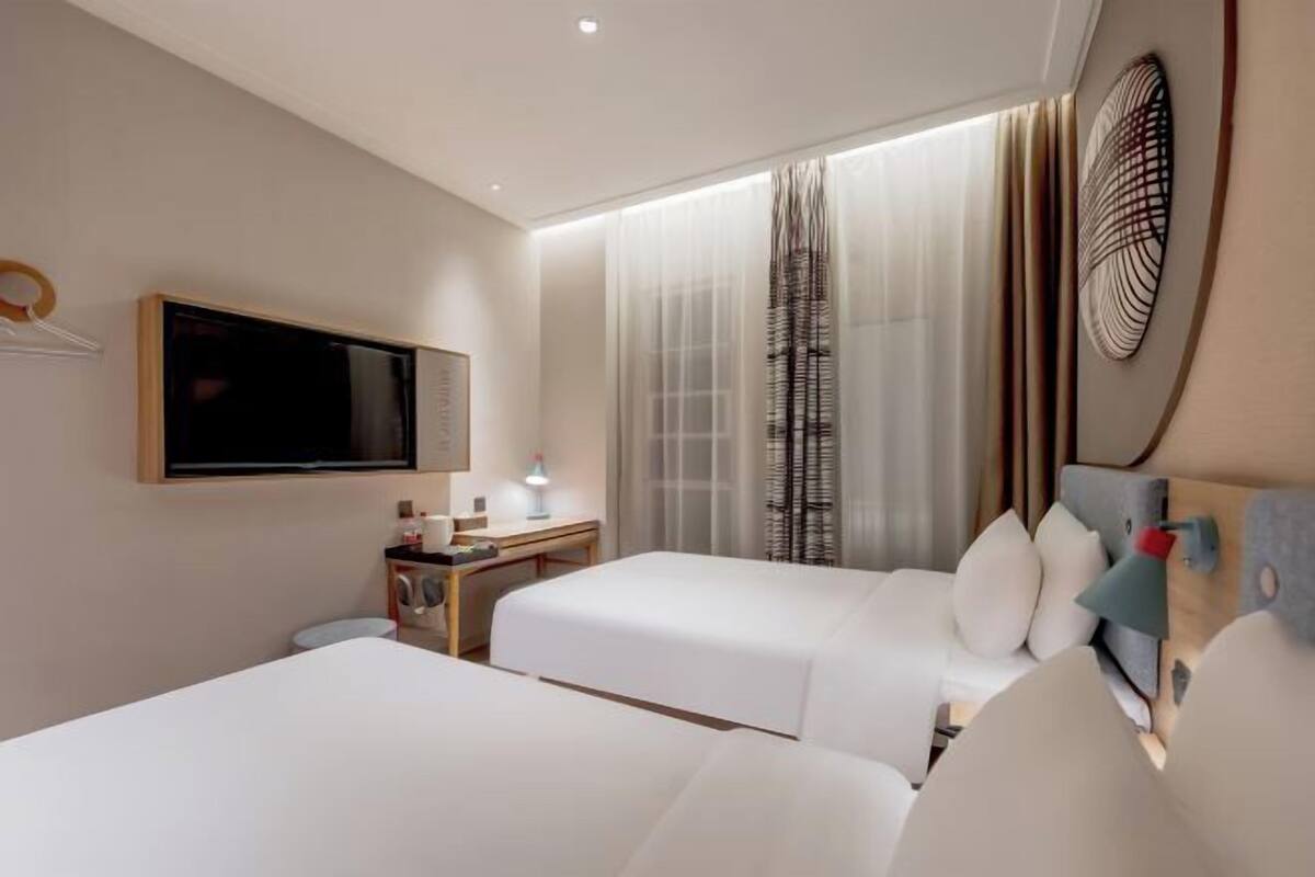 A hotel room with two beds, a TV, a desk, and a window with sheer curtains.