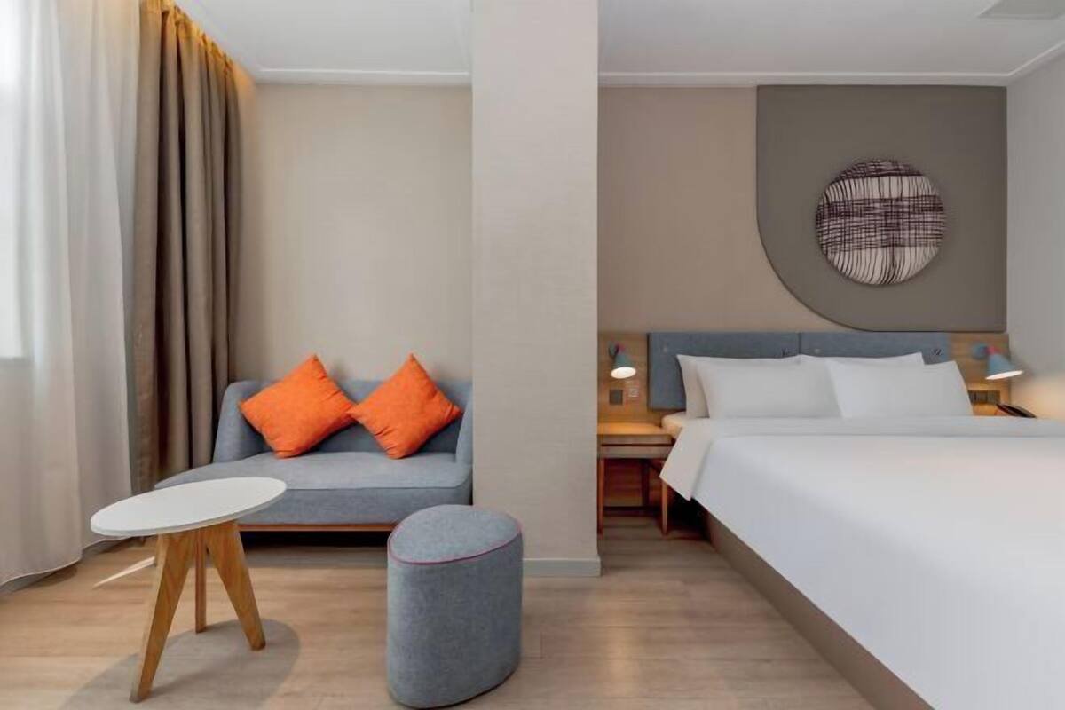 A hotel room with a bed, a sofa with orange pillows, a small round ottoman, a round wall decoration, and a nightstand with a lamp.