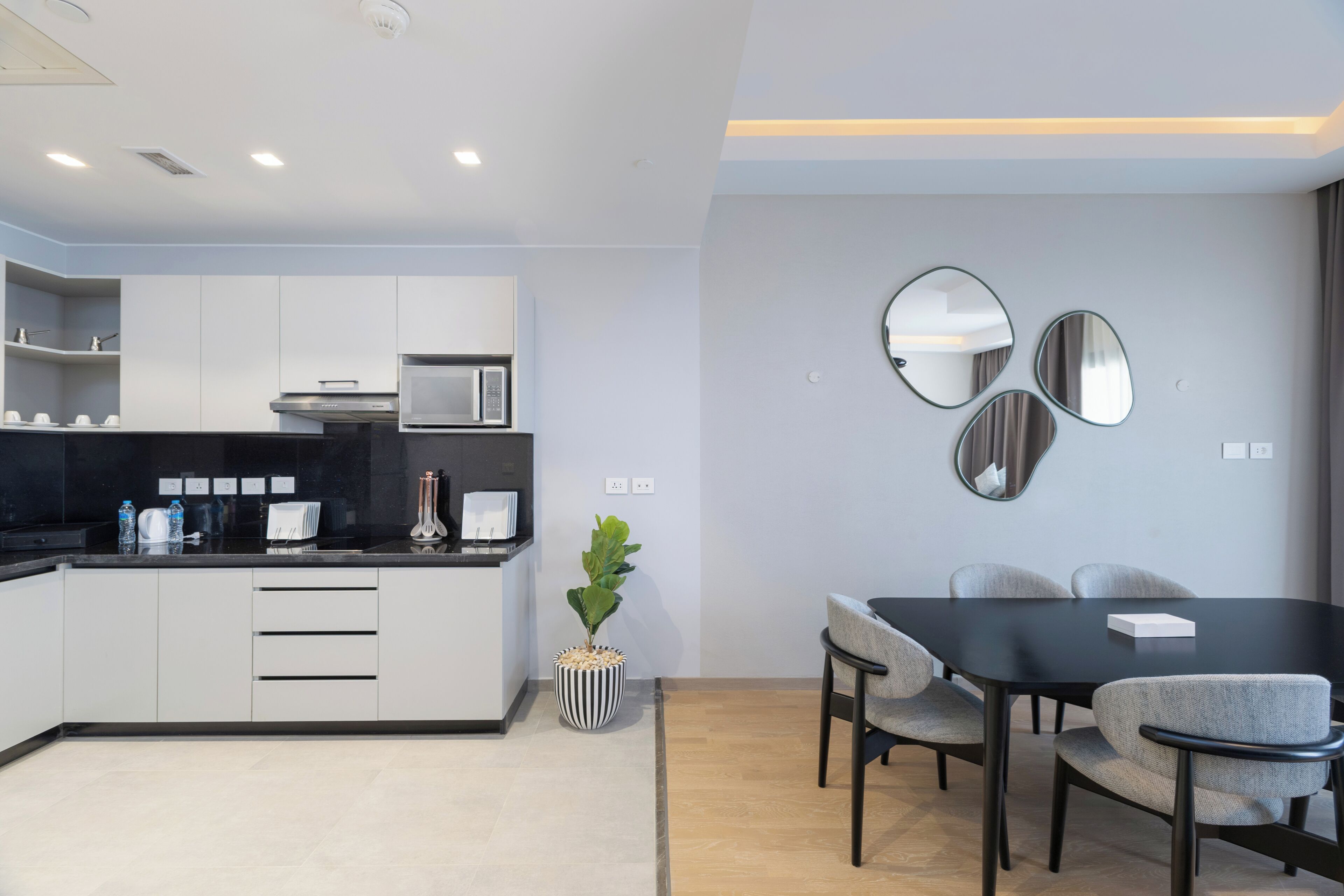 A modern kitchen with white cabinets, a black countertop, and a microwave. There is a dining table with chairs and a potted plant.