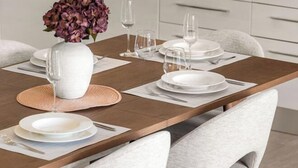 Una mesa de comedor dispuesta con platos blancos, cubiertos y un jarrón con flores moradas.