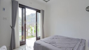 A bedroom with a bed, a sliding glass door leading to a balcony, and a view of greenery outside. - Batur Kencana Villa Mitra RedDoorz (Bali)