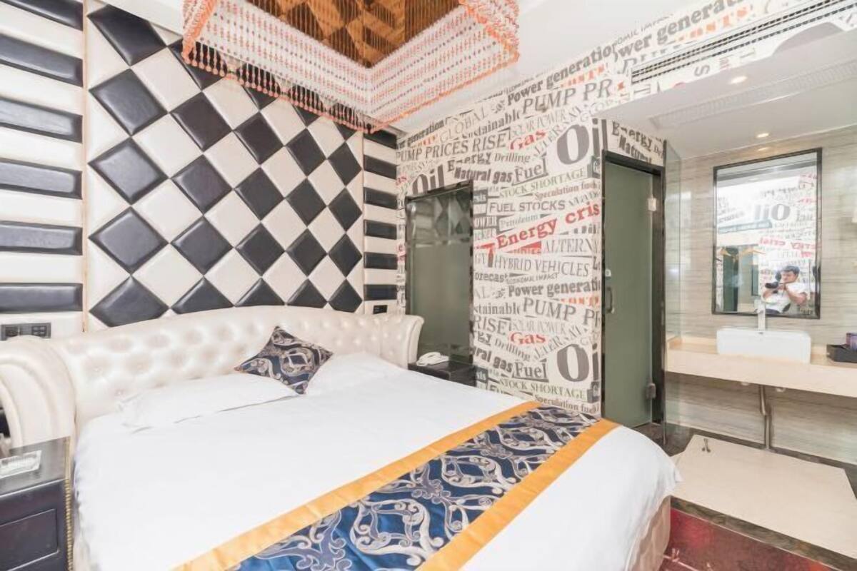 Una habitación de hotel moderna con cabecera a cuadros, cama con colcha estampada, un espejo grande y un baño con lavamanos y espejo.