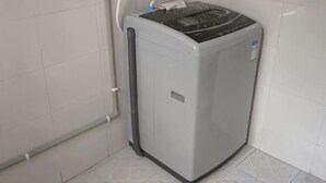 A compact, grey, wall-mounted laundry machine in a corner with a visible control panel and a small label on the side.