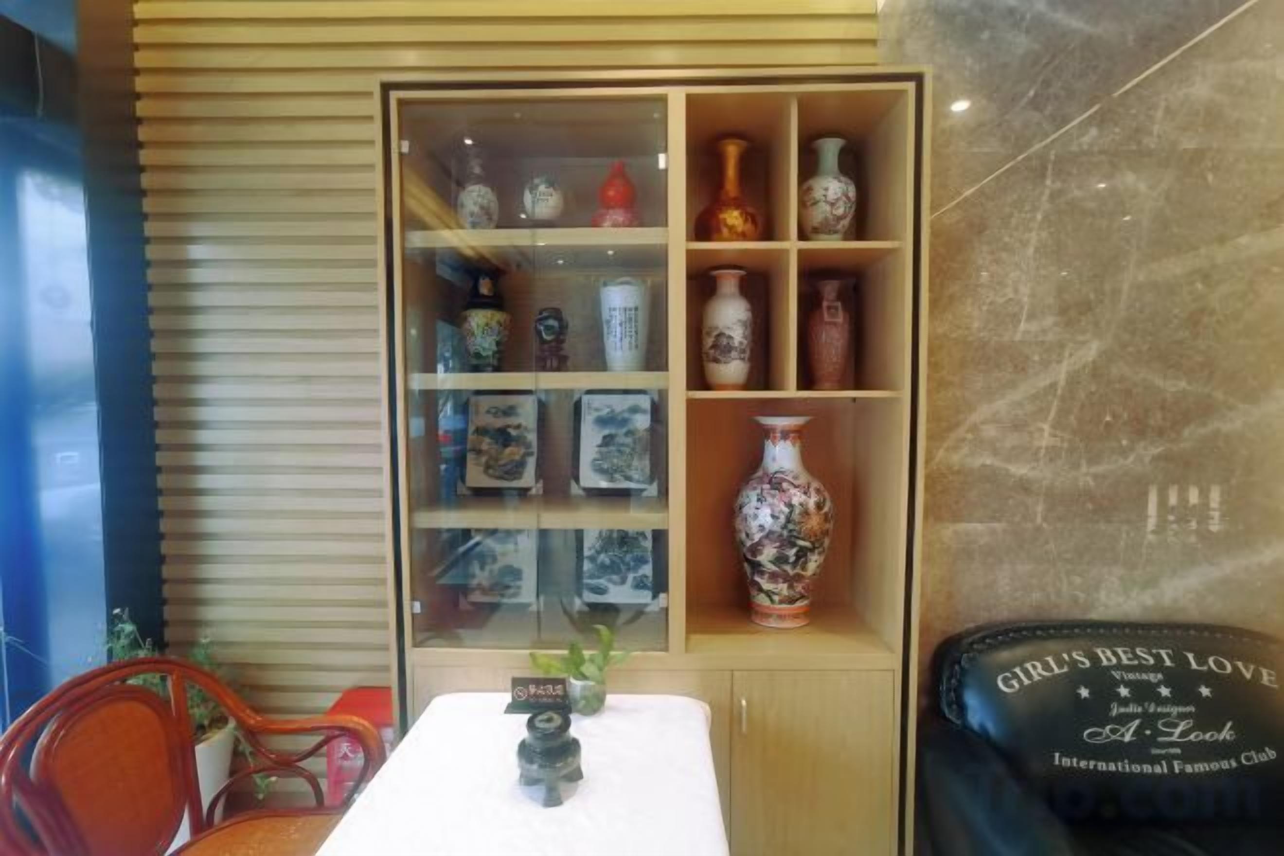 A room with a wooden slatted wall, a glass display case with decorative items, a table with a vase, and a chair.