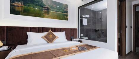 A hotel room with a large bed, a painting of a traditional building, a modern shower, and a large window.