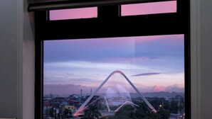 Una habitación con un amplio ventanal que ofrece vistas a un paisaje urbano al atardecer, destacando un puente y edificios urbanos.