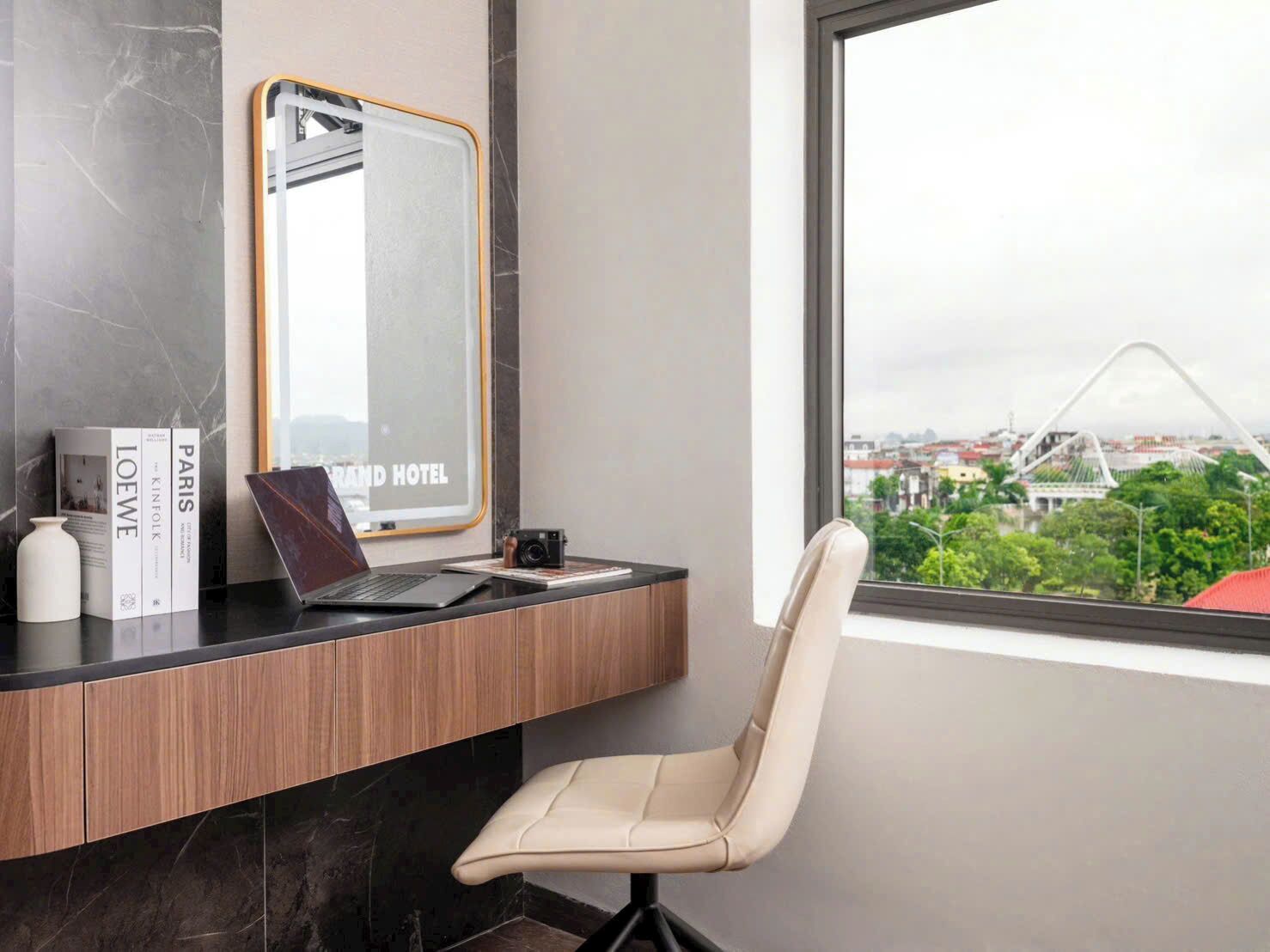 Una habitación de hotel moderna con un ordenador portátil sobre un escritorio de madera, una silla y vistas a un paisaje urbano a través de un ventanal grande.