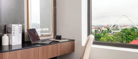 Habitació d'hotel moderna amb un ordinador portàtil sobre una taula de fusta, una cadira i vista a un paisatge urbà a través d'una finestra gran.