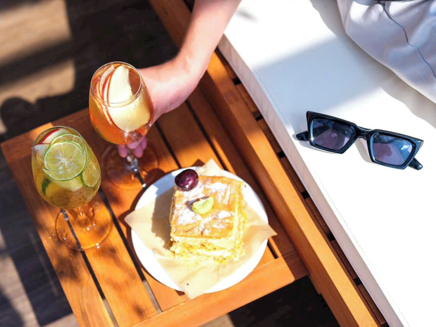 A wooden table with a plate of food, a drink, and sunglasses.