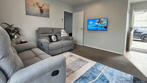 A living room with a gray sectional sofa, a flat-screen TV on the wall, a painting of a deer, and a door leading outside. -  Kozy Kottage, Near Hospital & Perfect for Families  (Hamilton)