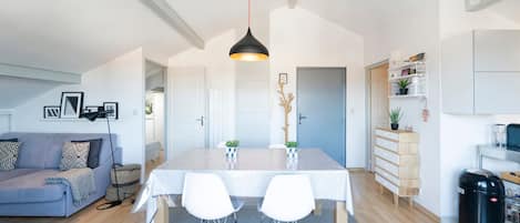 A modern kitchen with a dining table, chairs, a hanging light fixture, a sofa, and a small plant.