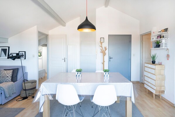 Een moderne woonkamer met een eettafel, witte stoelen, een zwart hanglamp, een grijze bank en een houten kast met planten.