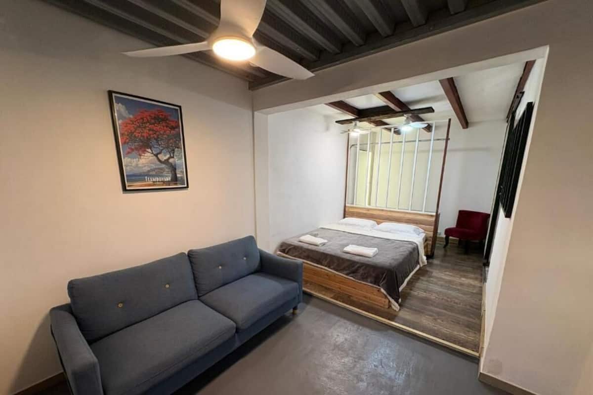 A room with a bed, a blue sofa, a ceiling fan, a framed picture on the wall, and a window with blinds.