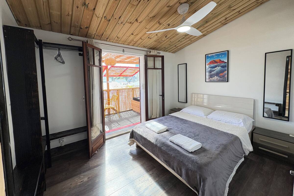 A bedroom with a wooden ceiling, a bed with a grey bedspread, a white headboard, a bedside table, and a large mirror.