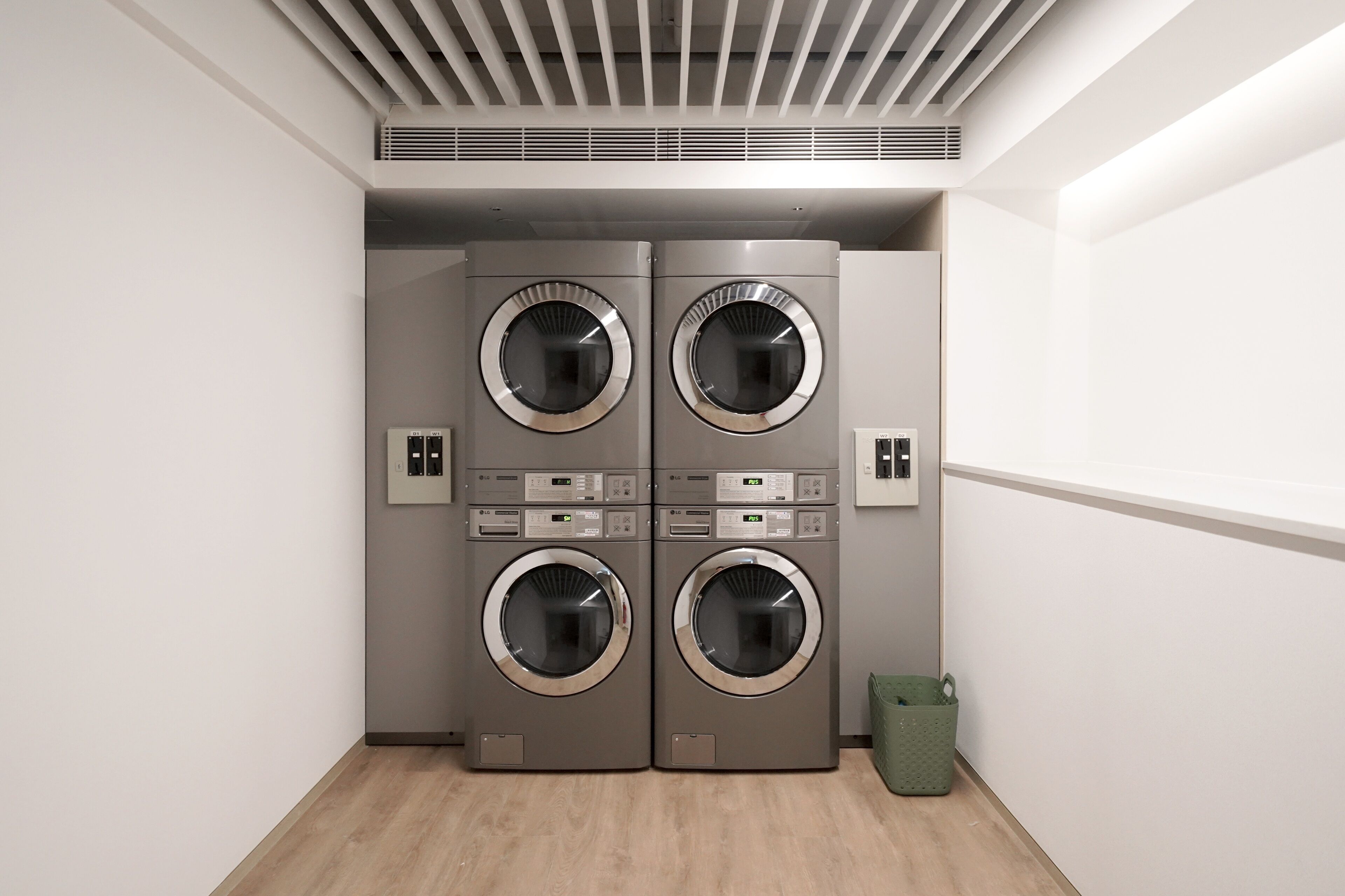 A laundry room with four wall-mounted washing machines, a green waste basket, and a vent on the ceiling.
