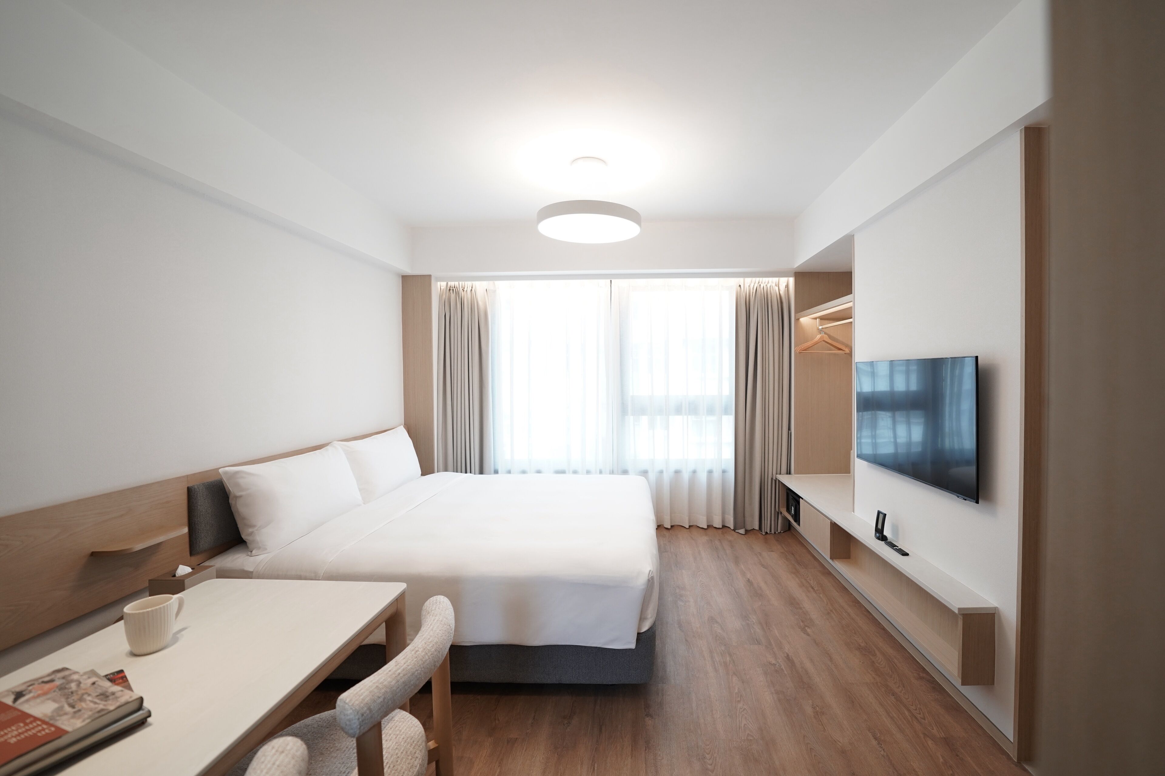 A modern hotel room with a large bed, a desk with a chair, a flat-screen TV mounted on the wall, and a wardrobe.