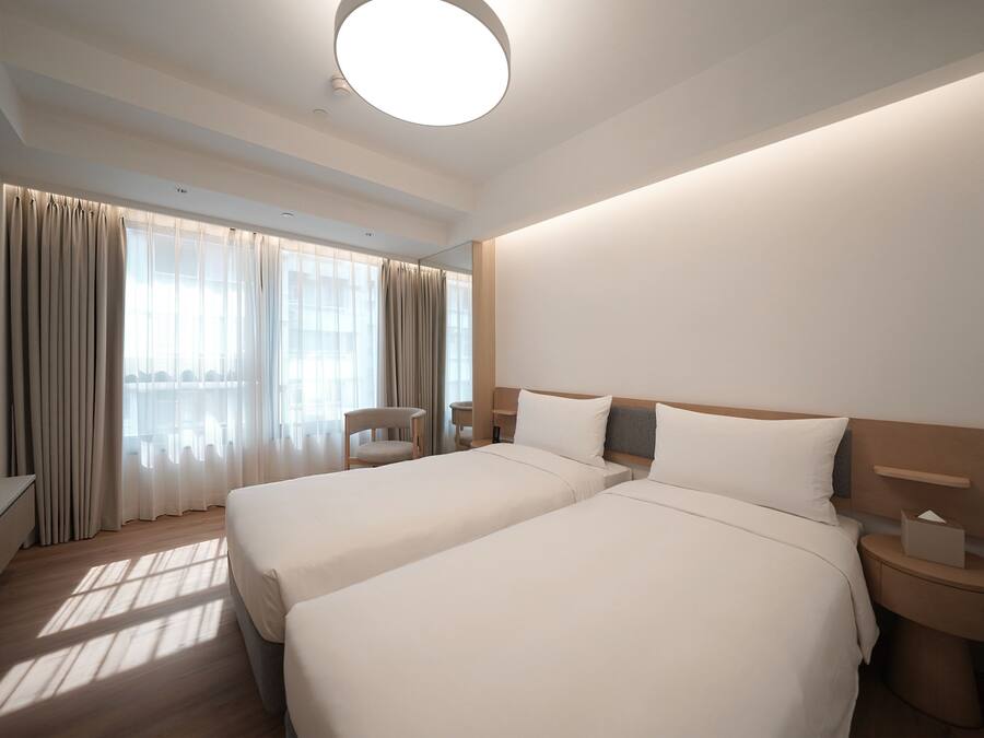 Habitación de hotel con dos camas, un escritorio, una silla y un ventanal con cortinas.