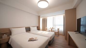 A hotel room with two beds, a desk, a TV, and a window with curtains.