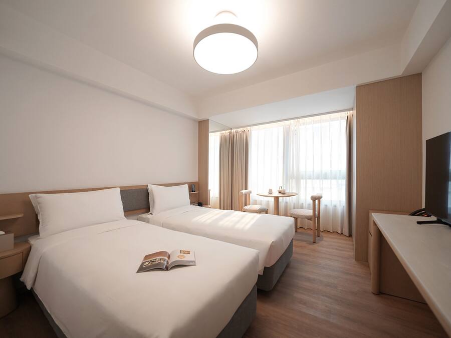 A hotel room with two beds, a desk, a TV, and a window with curtains.