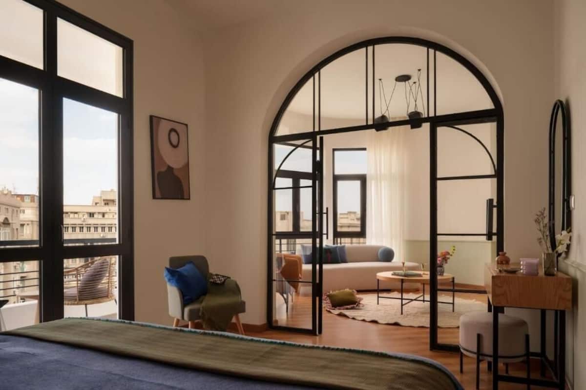 Une chambre d’hôtel moderne dotée d’une grande fenêtre en arc de cercle, d’un lit, d’un coin salon avec un canapé et des fauteuils, et d’une petite table.