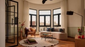 Un salon moderne avec un canapé, un fauteuil et une table basse, de grandes fenêtres offrant une vue sur la ville et une porte vitrée donnant sur un espace extérieur.