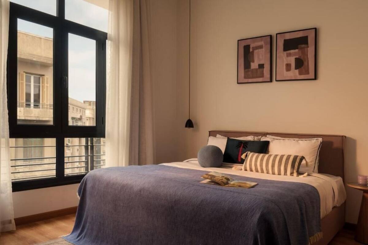 Une chambre avec une grande fenêtre, un lit recouvert d’une couverture bleue et deux œuvres d’art encadrées accrochées au mur.