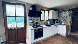 A compact kitchen with a gas stove, microwave, and built-in oven. A stone wall and a clock are visible in the background. - Gîte 6 people 15min from Angers (Loire-Authion)