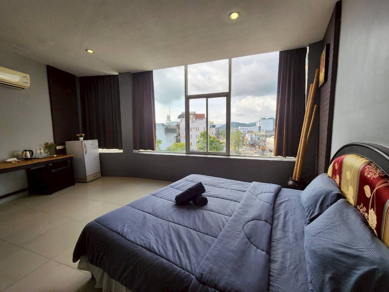 A hotel room with a large bed, a desk, a refrigerator, and a window with a city view.