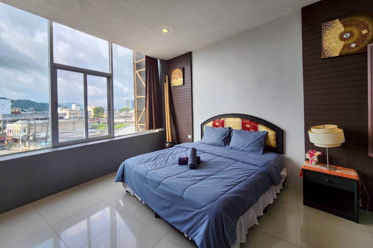 A modern hotel room with a large bed, a nightstand, a window with a city view, and a decorative wall piece.