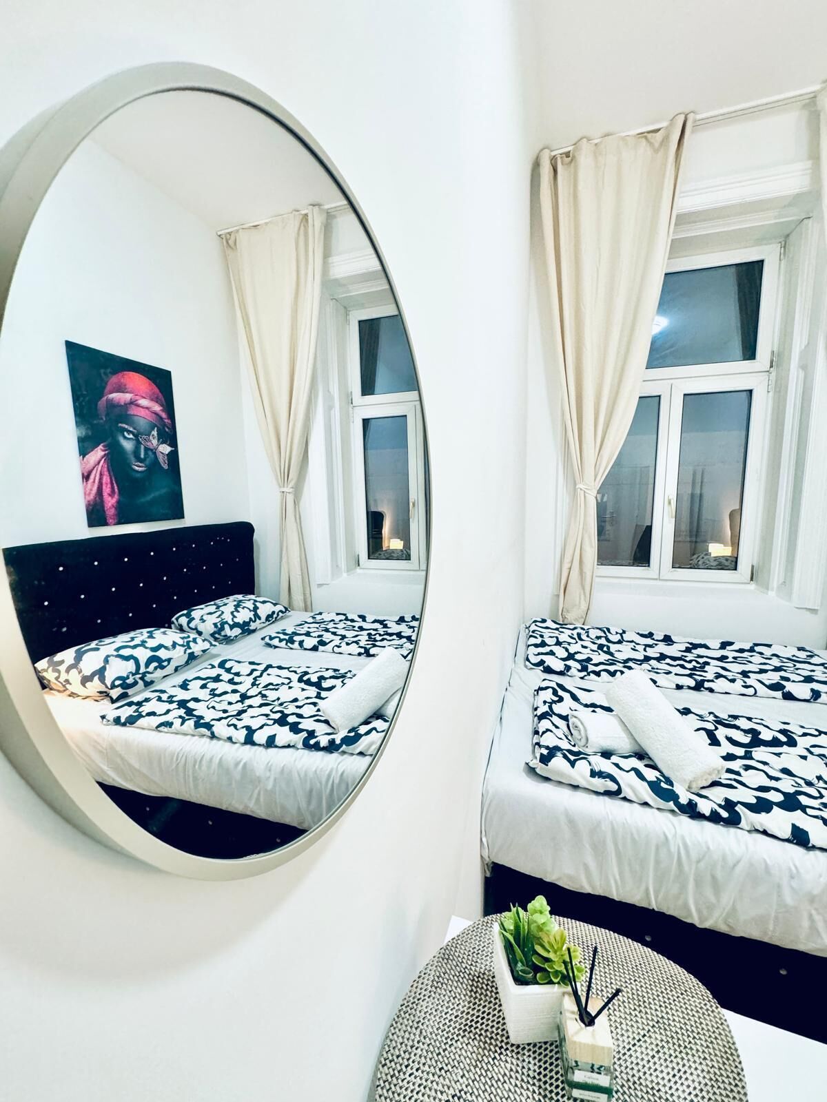 Habitación con espejos, dos camas, un espejo redondo, una ventana con cortinas y una pequeña planta sobre una mesa redonda.