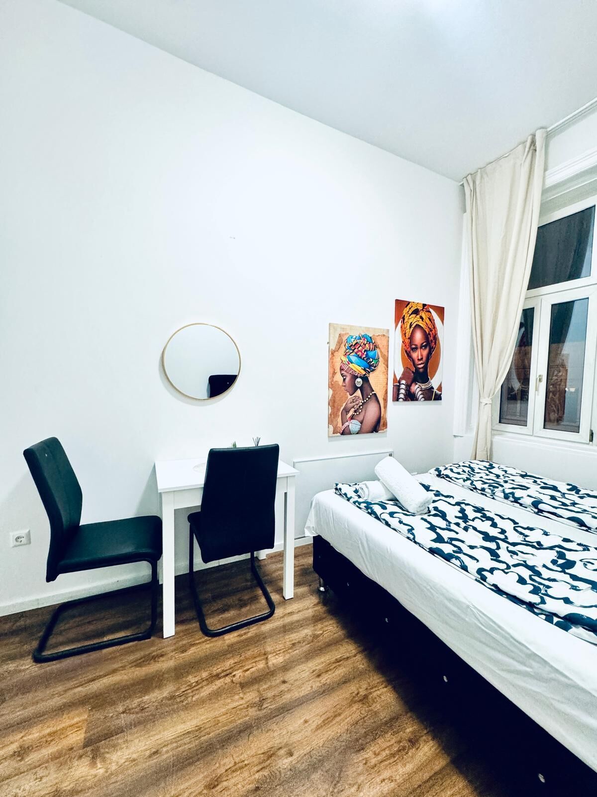 A bedroom with a bed, a desk, two chairs, a round mirror, and two wall art pieces.