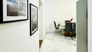 Un pasillo con paredes blancas, suelo de mármol y cuadros en la pared. Se observa un comedor con mesa y sillas al fondo.