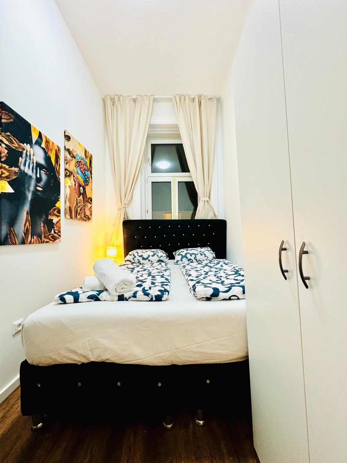 A bedroom with a bed, a window with curtains, a nightstand, and wall art.