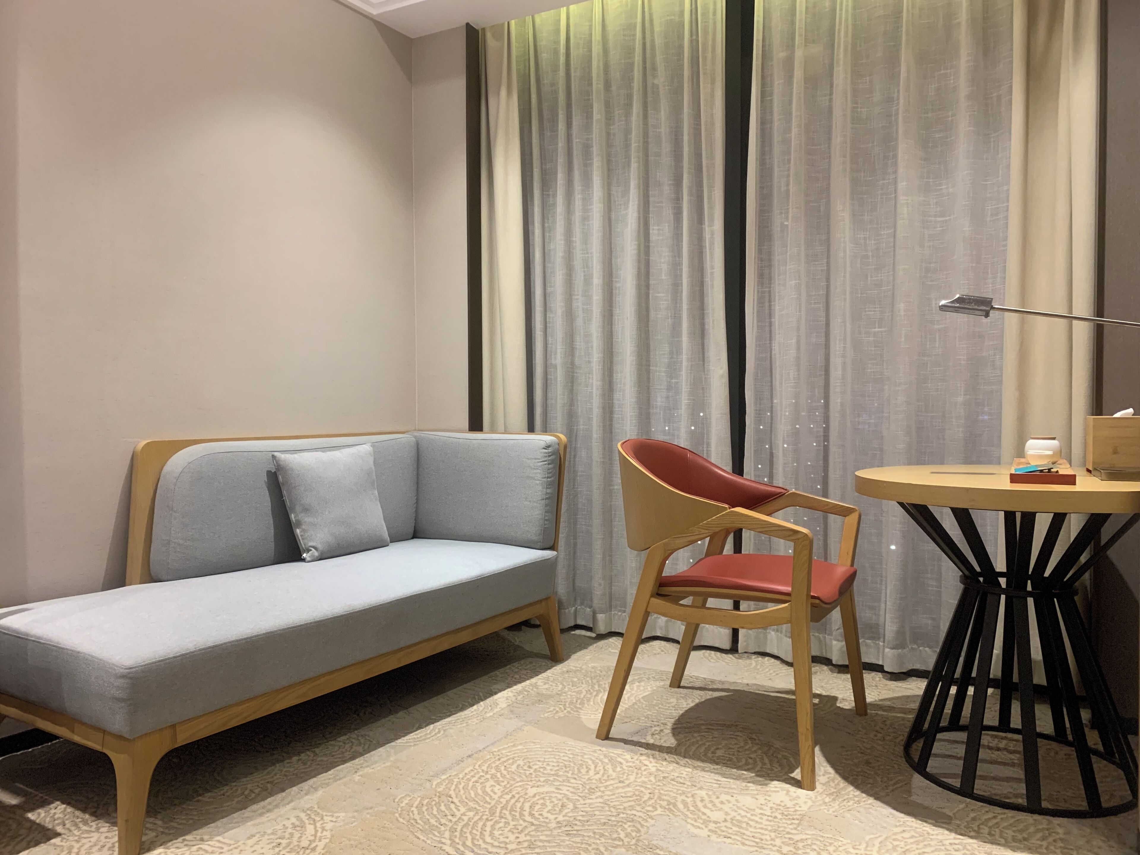 Ein Zimmer mit einem hellgrauen Sofa, einem Holzstuhl, einem kleinen runden Tisch mit einer Lampe und einem gemusterten Teppich.