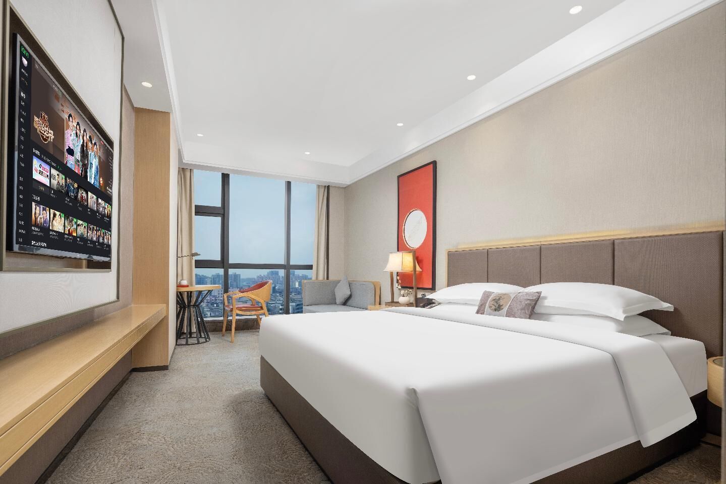Een hotelkamer met een groot bed, een bureau, een stoel, een aan de muur bevestigde televisie en een groot raam met uitzicht op de stad.