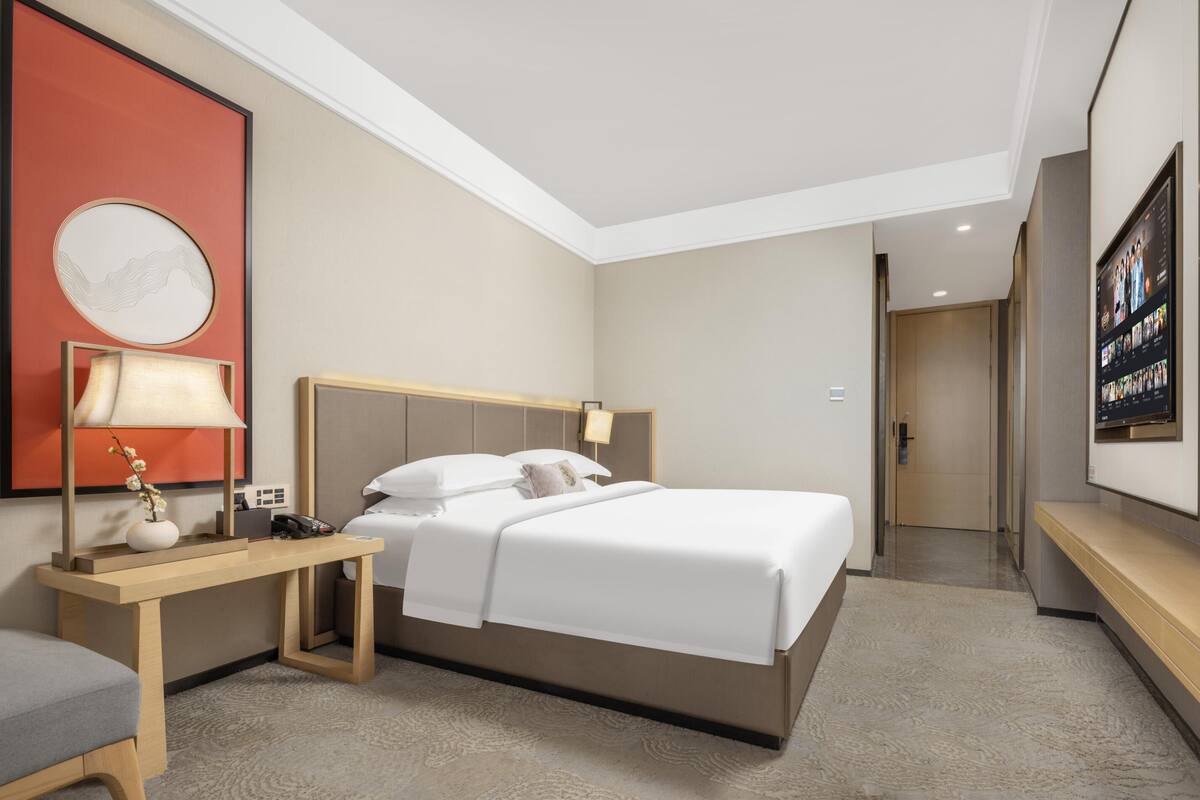 Ein Hotelzimmer mit Bett, Nachttisch, Lampe und einem großen gerahmten Kunstwerk an der Wand.