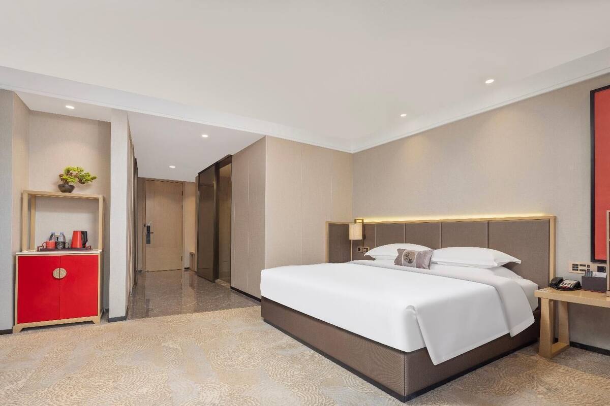 Ein Hotelzimmer mit einem großen Bett, einer roten Minibar, einem Schreibtisch mit Lampe sowie einer Badezimmertür.
