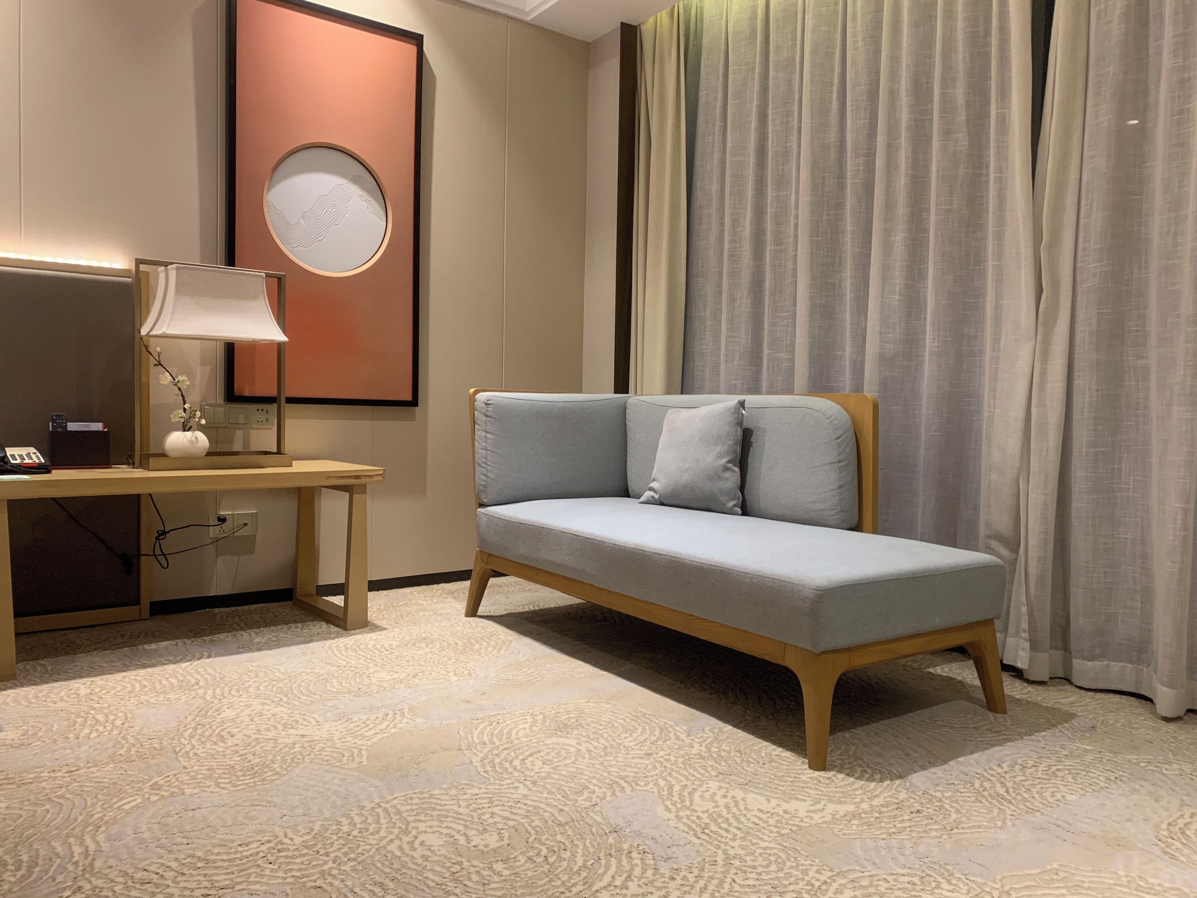 A hotel room with a light blue sofa, a wooden desk with a lamp, a framed picture on the wall, and a large window with curtains.