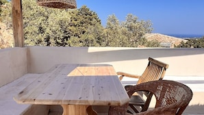 A wooden table and wicker chairs under a wooden pergola with a hanging light fixture, set against a clear blue sky and distant landscape. - DomoMea Porto Cervo (Arzachena)