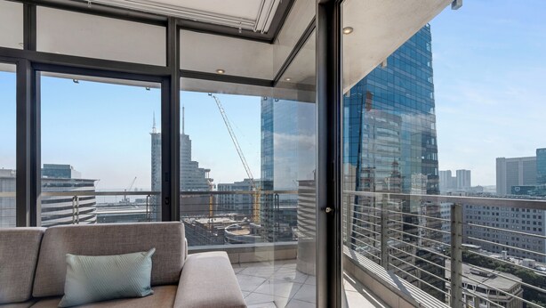 A modern balcony with a sofa and a glass door, offering a city view with high-rise buildings and construction cranes. - Modern apartment with skyline and sea views (Cape Town)
