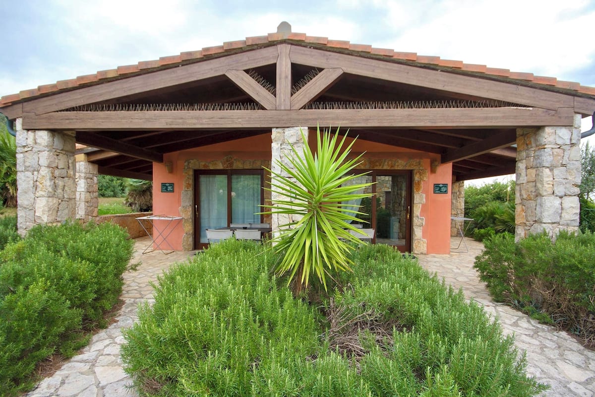 A stone building with a wooden roof, surrounded by green shrubs and a palm tree.