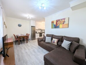 A living room with a brown sofa, a dining table with chairs, a kitchen area, and a clock on the wall.