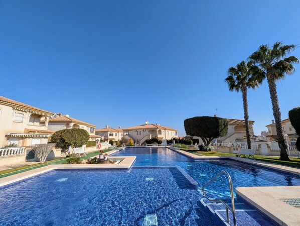 A swimming pool with a tiled deck, surrounded by buildings with Mediterranean architecture, palm trees, and a clear blue sky.