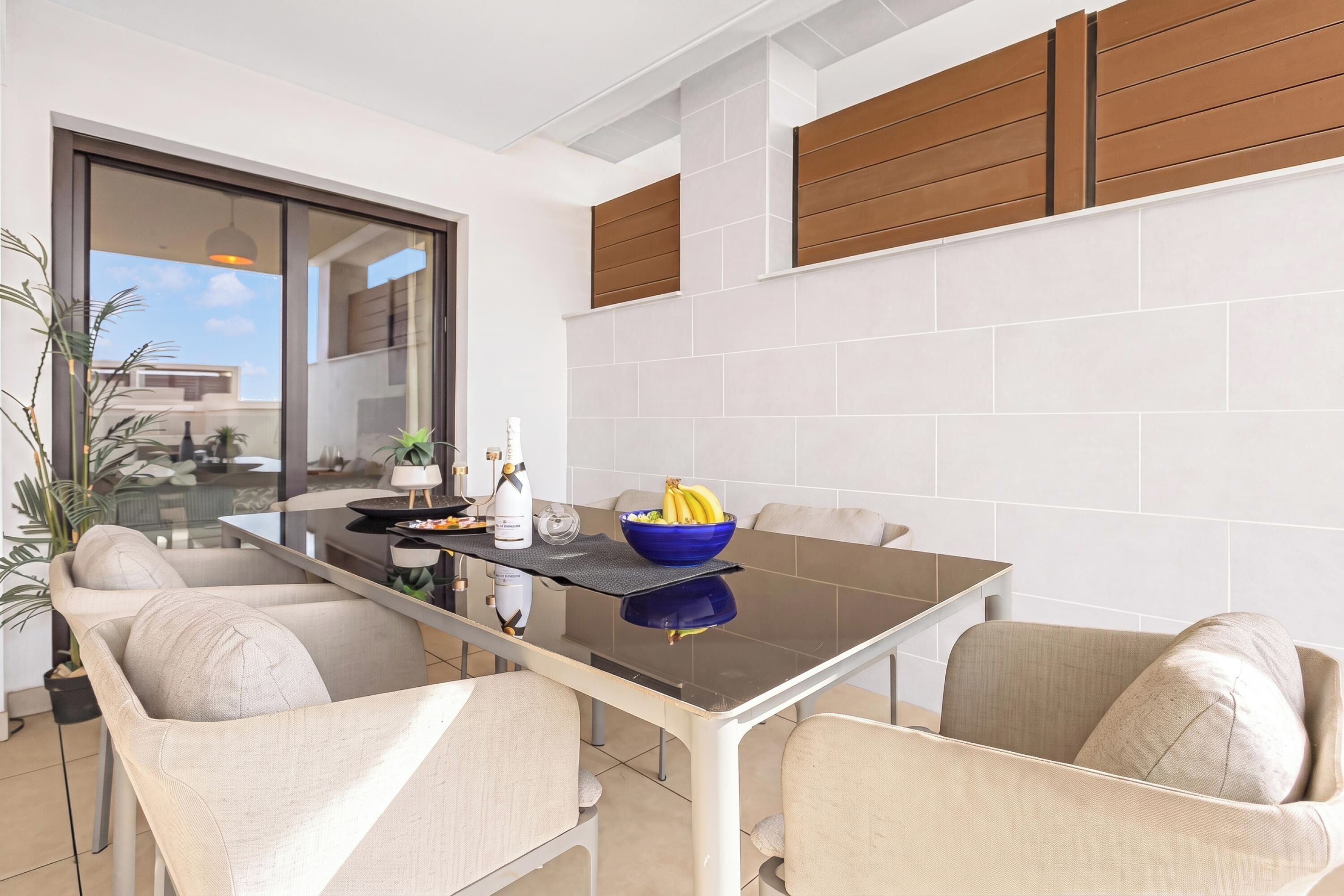 A modern dining area with a glass-top table, four chairs, a blue bowl with fruit, and a bottle of champagne.