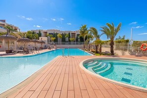 Una piscina con muelle de madera, sillas de descanso, palmeras y un edificio al fondo.
