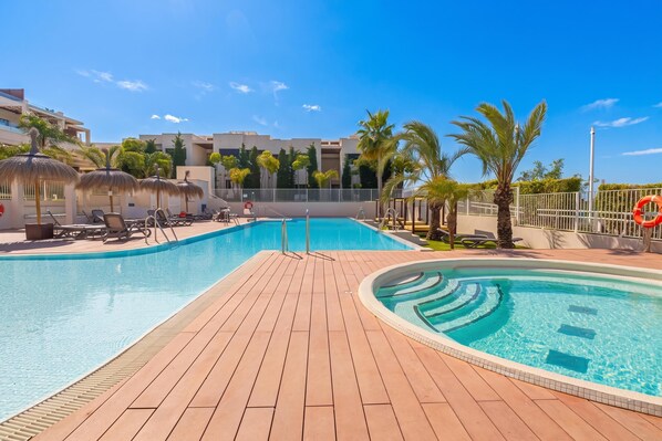 A swimming pool with a wooden deck, lounge chairs, palm trees, and a building in the background. - Lotus Jardinana Paradise (Mijas)