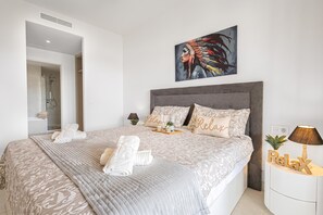 A modern bedroom with a large bed, a headboard, a painting, a bedside table with a lamp, and a wall-mounted light fixture. - Lotus Jardinana Paradise (Mijas)